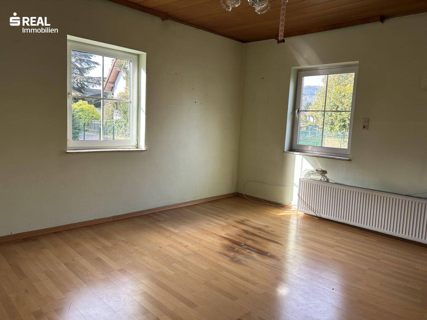 Heller Wohnraum mit Holzdecke, Parkettboden und zwei Fenstern mit Blick ins Grüne.