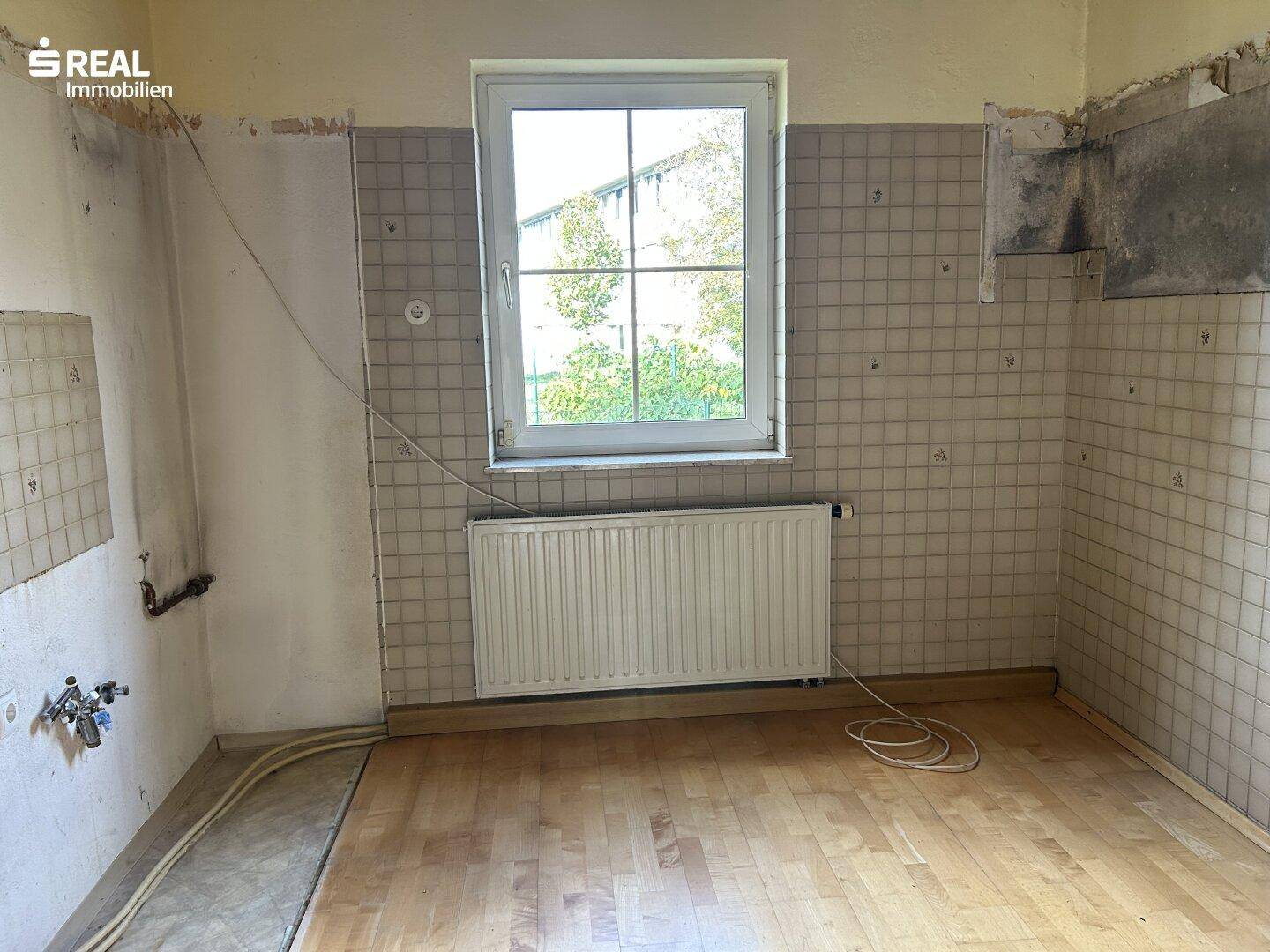 Leere Küche mit teilweise gefliesten Wänden, Heizkörper und Fenster mit Blick ins Grüne.