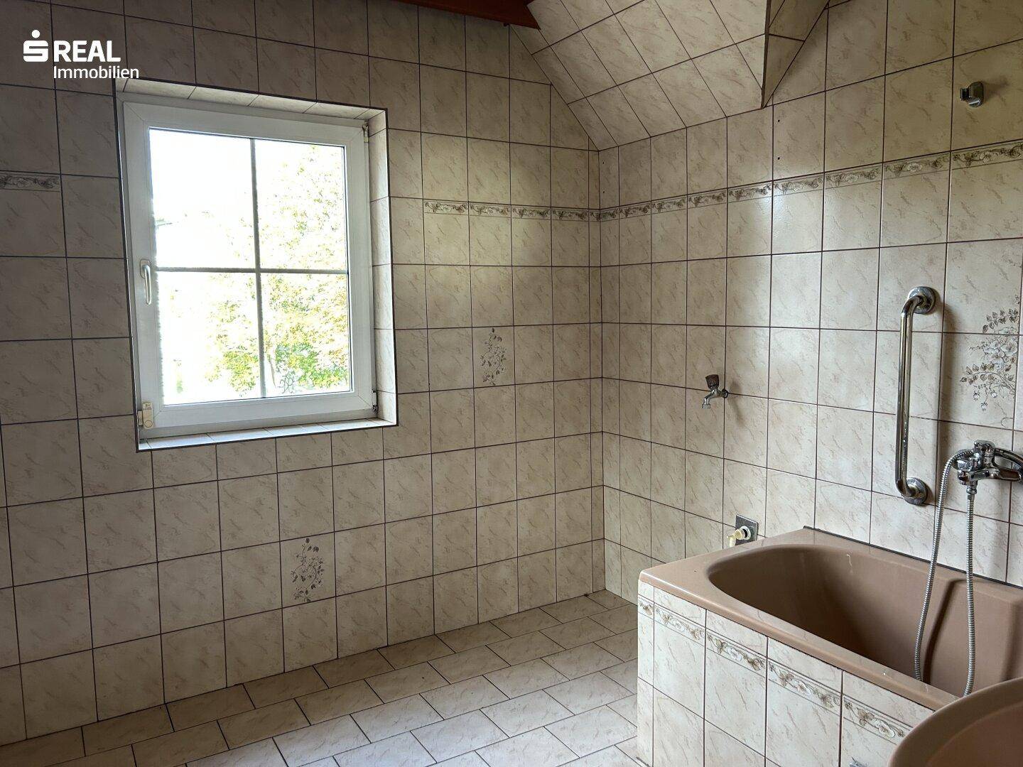 Badezimmer mit beigefarbenen Fliesen, Badewanne, Haltegriffen und Fenster für Tageslicht.