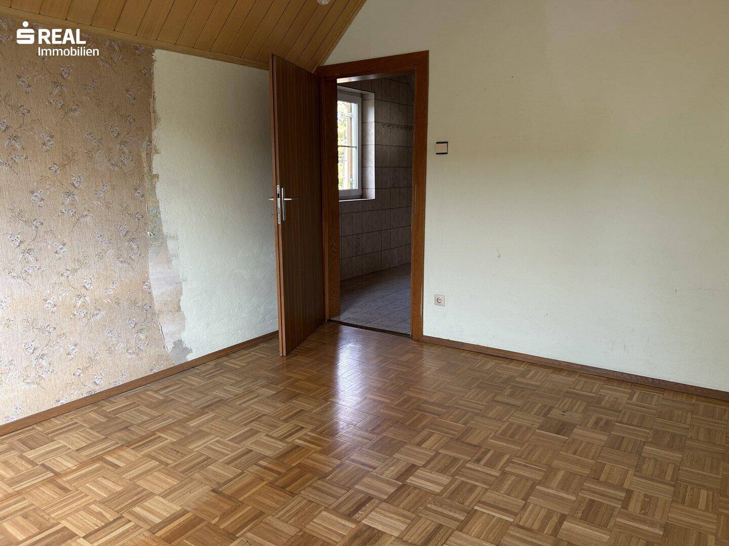 Wohnraum mit Parkettboden, Holzdecke und offener Tür zu einem gefliesten Nebenraum.