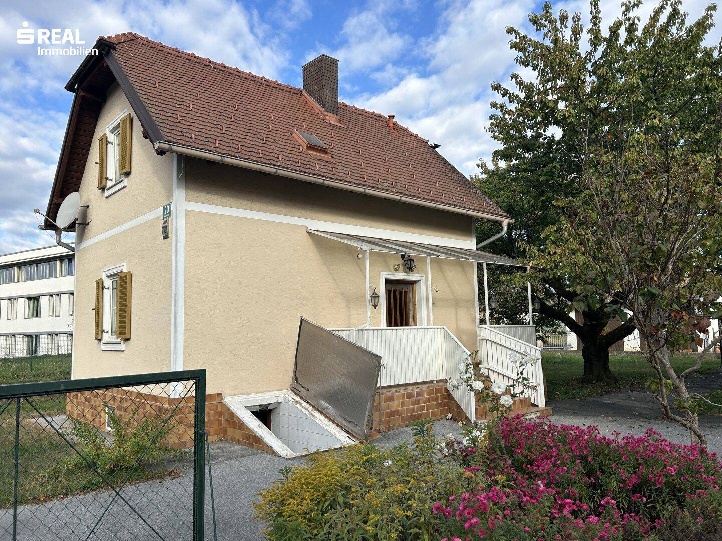 Hausfassade mit überdachtem Eingangsbereich, Kellerzugang und blühenden Sträuchern im Vorgarten.