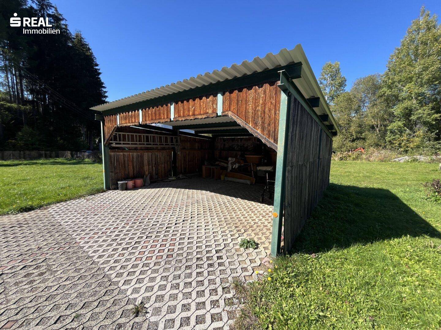 Offener Holzschuppen oder Carport mit Wellblechdach auf gepflastertem Untergrund im Grünen.