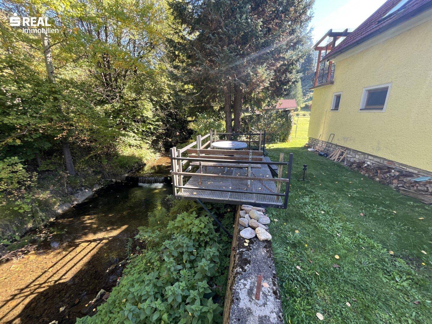 Holzterrasse mit Geländer über einem kleinen Bach, umgeben von üppiger Vegetation und Bäumen.