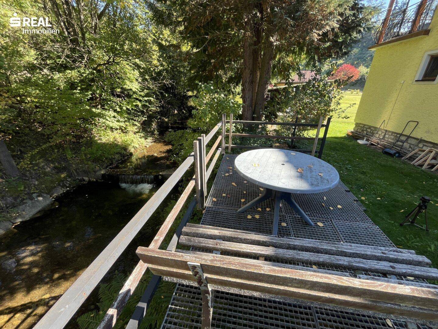 Kleine Terrasse mit Tisch und Stühlen direkt über einem Bachlauf, umgeben von Bäumen.