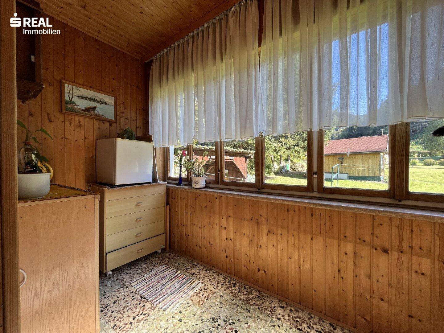 Heller, holzvertäfelter Wintergarten mit vielen Fenstern und Blick ins Grüne, gemütlich eingerichtet.