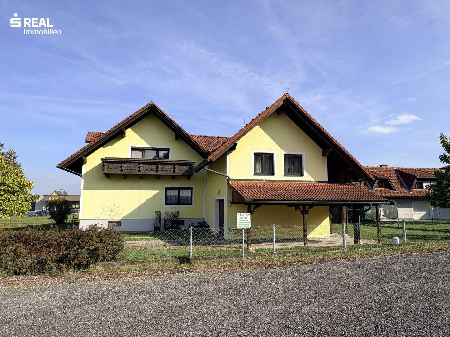 Vorderansicht des gelben Hauses mit rotem Ziegeldach, Balkon und überdachtem Eingangsbereich.