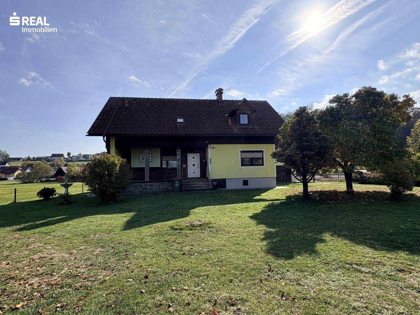 Frontansicht des Hauses mit großem, sonnigen Garten und altem Baumbestand unter blauem Himmel.