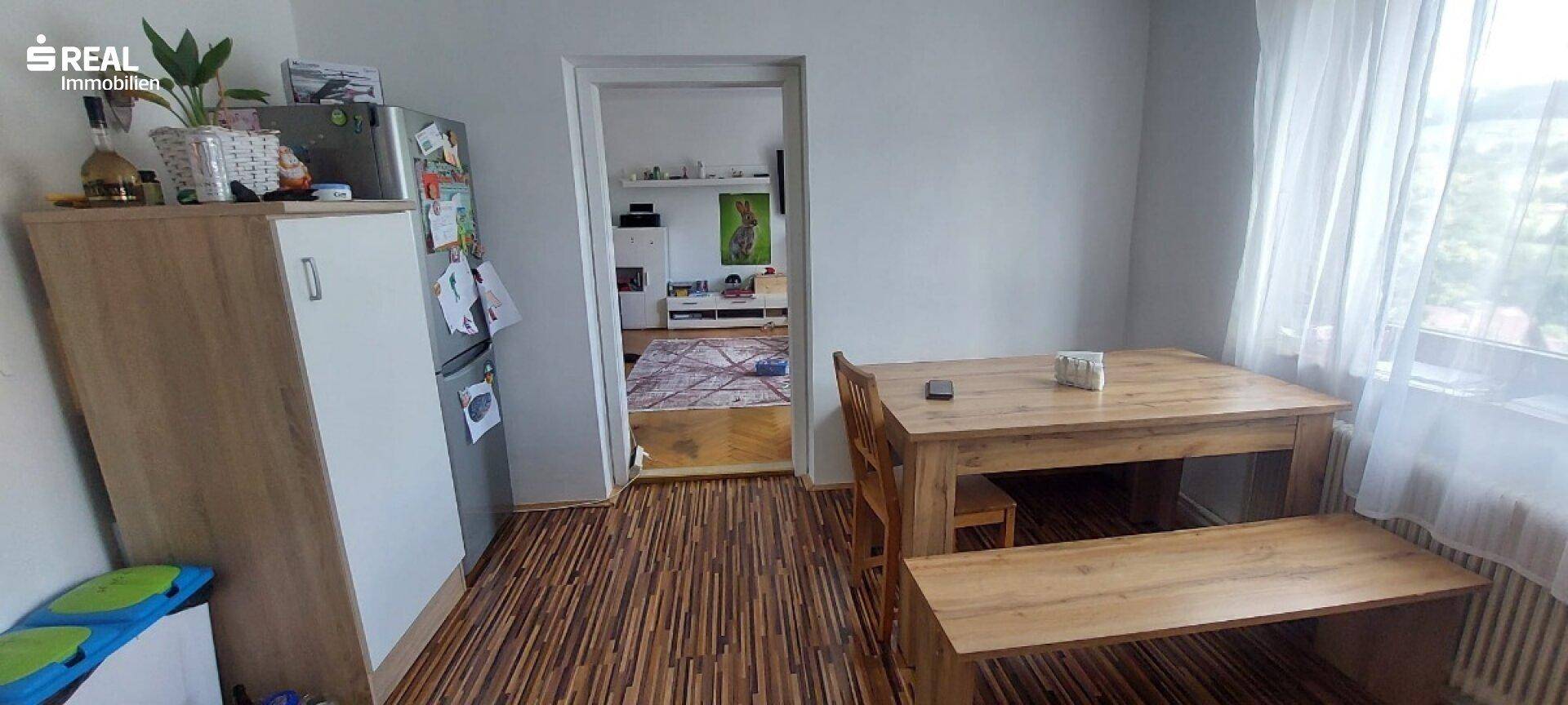 Essbereich mit Holztisch und Bank, angrenzend an einen weiteren Raum, mit Blick ins Grüne.