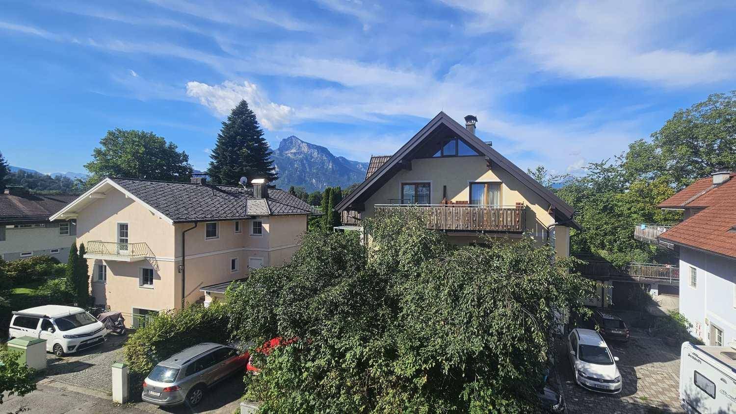 Häuser mit traditionellen Dächern und Balkonen, umgeben von Bäumen mit Bergblick.