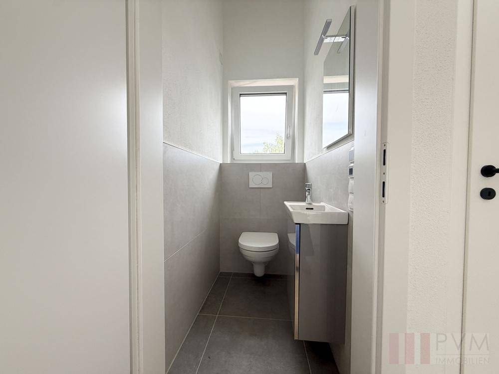 Modernes Gäste-WC mit Fenster, Wand-WC und kleinem Waschtisch in neutralen Farben.