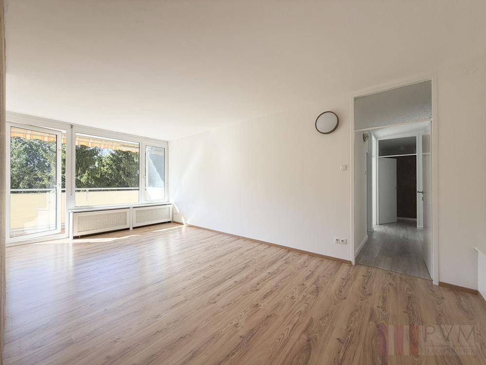 Leeres Wohnzimmer mit Laminatboden, großen Fenstern und Zugang zu einem Balkon.