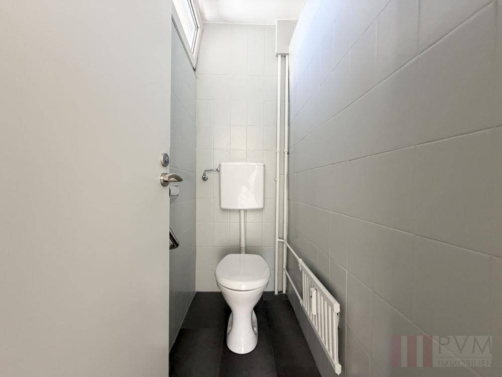 Separates WC mit grauen Fliesen, wandmontierter Toilette und kleinem Fenster.