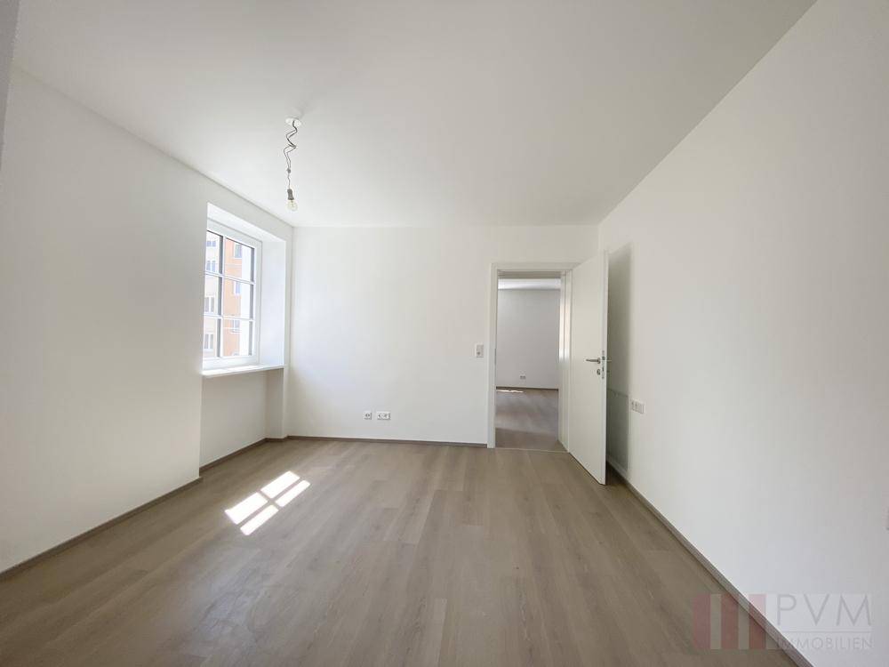 Lichtdurchfluteter Raum mit hellem Laminatboden und Zugang zu einem angrenzenden Zimmer.