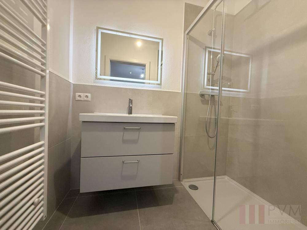 Modernes Badezimmer mit beleuchtetem Spiegel, Waschtisch und ebenerdiger Dusche.