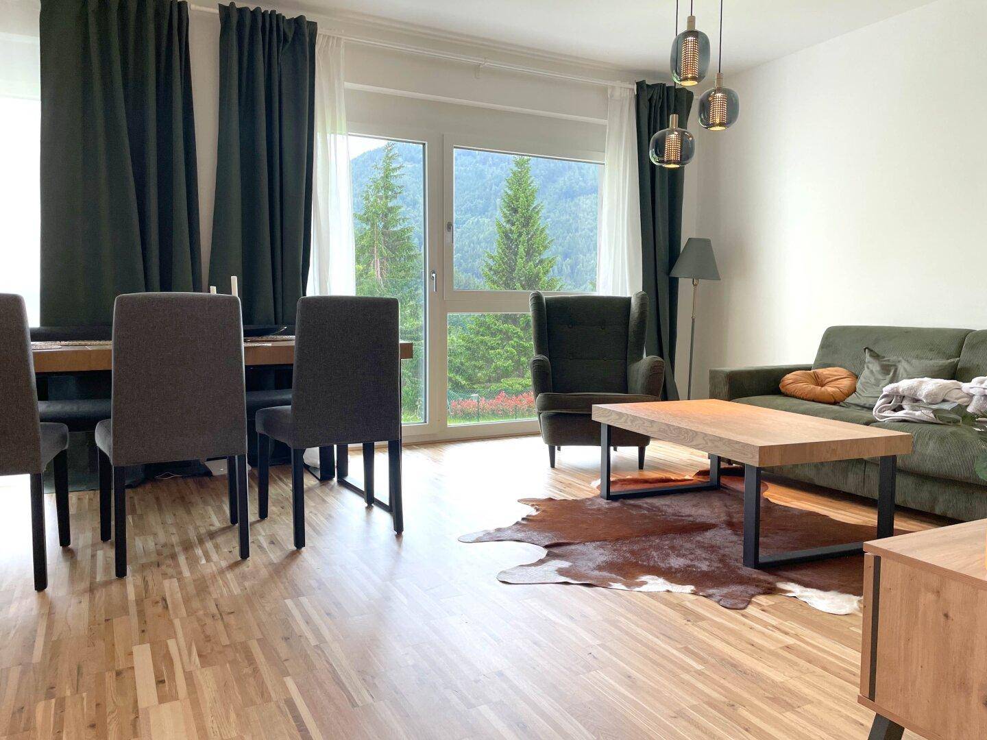 Gemütlicher Wohnbereich mit Holzboden, grüner Couch und großem Fenster mit Bergblick.