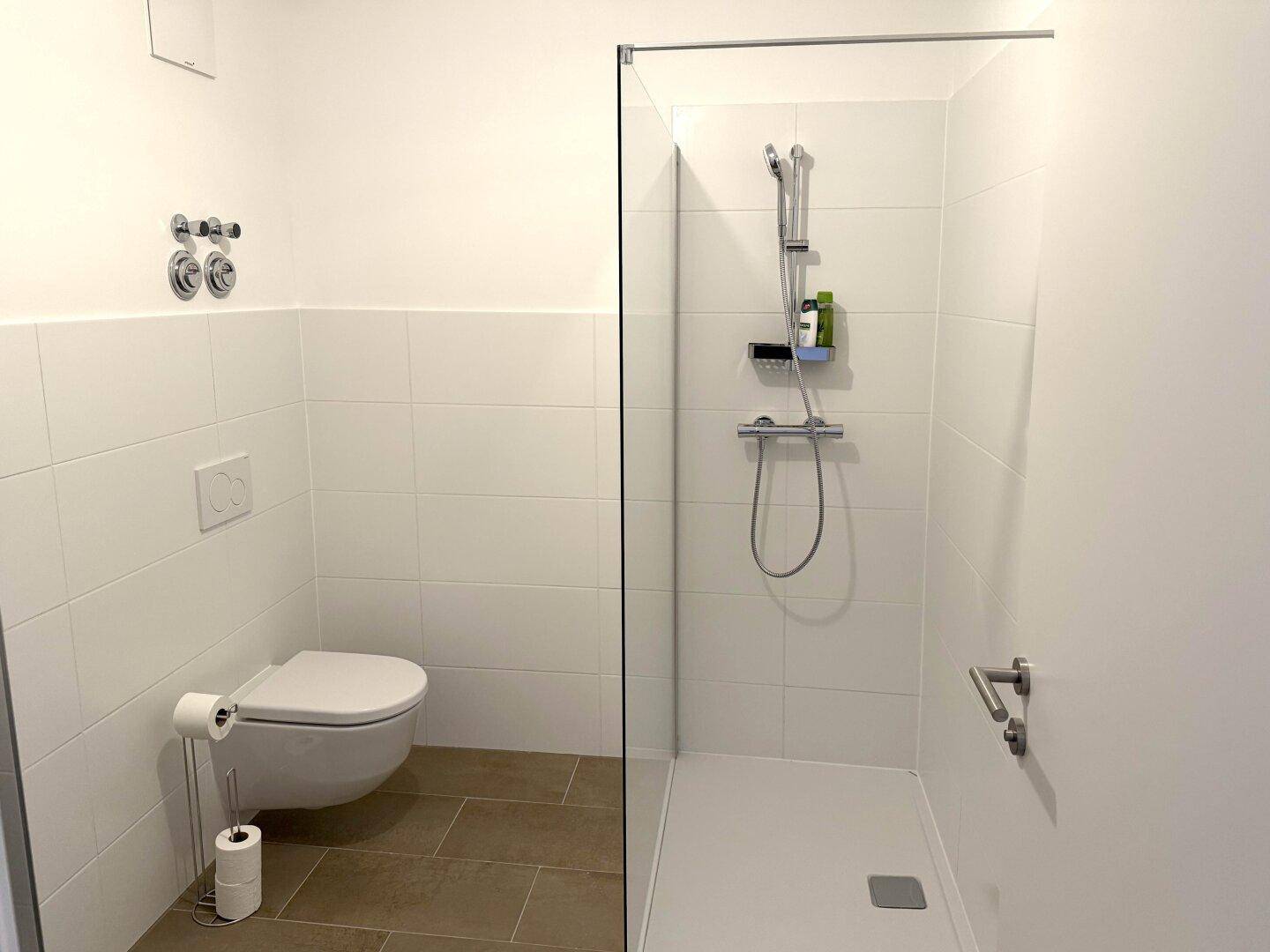 Helles Badezimmer mit weiß gefliesten Wänden, braunem Boden und einer modernen Duschkabine.