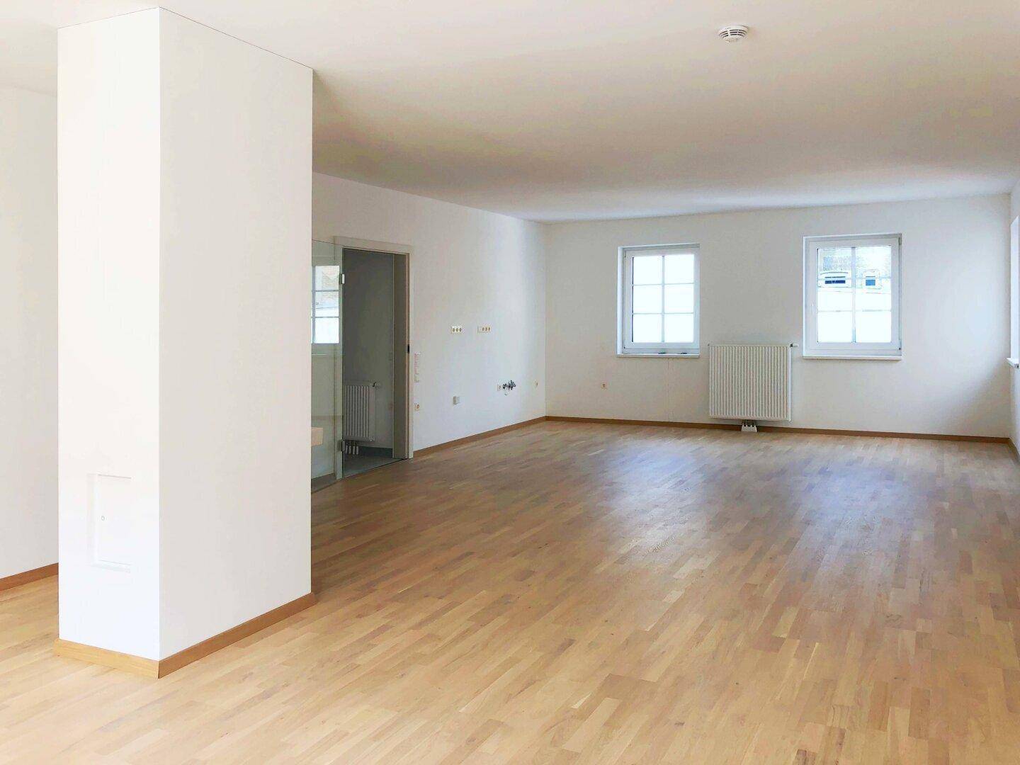 Geräumiger, leerer Raum mit hellem Parkettboden, großen Fenstern und viel Tageslicht.