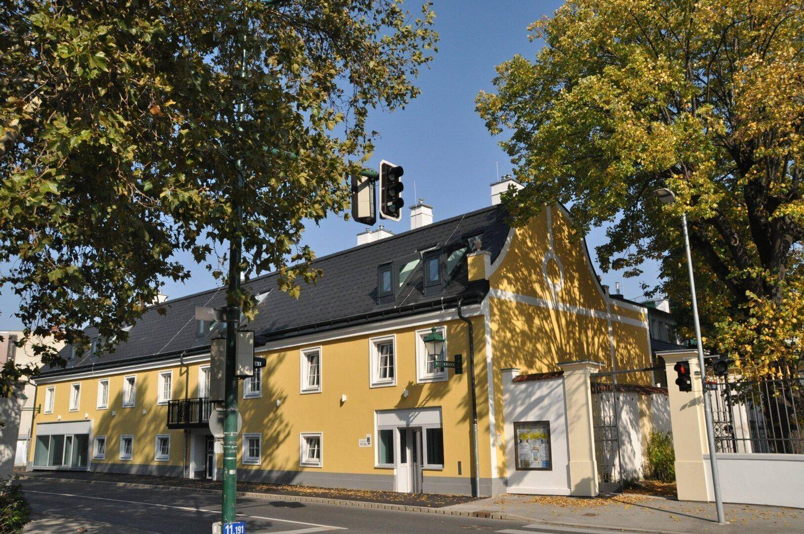 Historisches gelbes Gebäude an einer Straße, umgeben von Bäumen unter blauem Himmel.