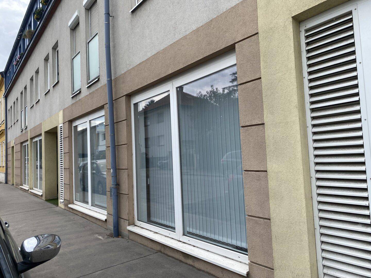 Fassade eines Geschäftsgebäudes mit großen Schaufenstern, Jalousien und einem Gehweg davor.