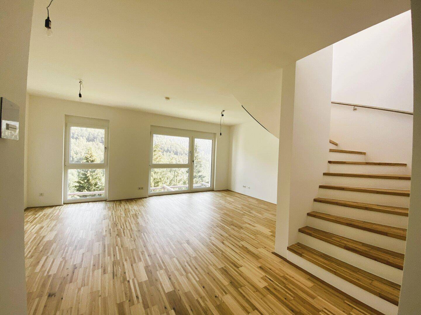 Heller Wohnbereich mit Holzparkettboden, großen Fenstern und offener Treppe.