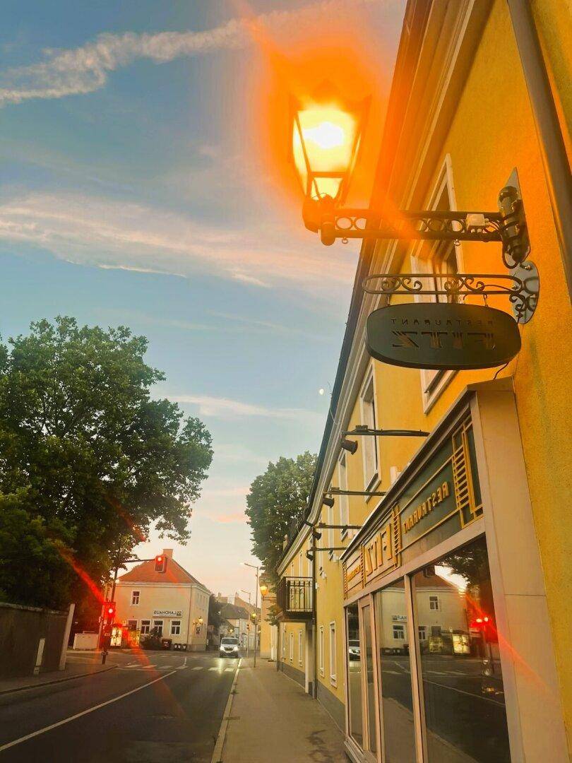 Gelbes Gebäude mit leuchtender Laterne und Geschäftsauslage an einer ruhigen Straße bei Abenddämmerung, mit Bäumen und weiteren Häusern im Hintergrund.