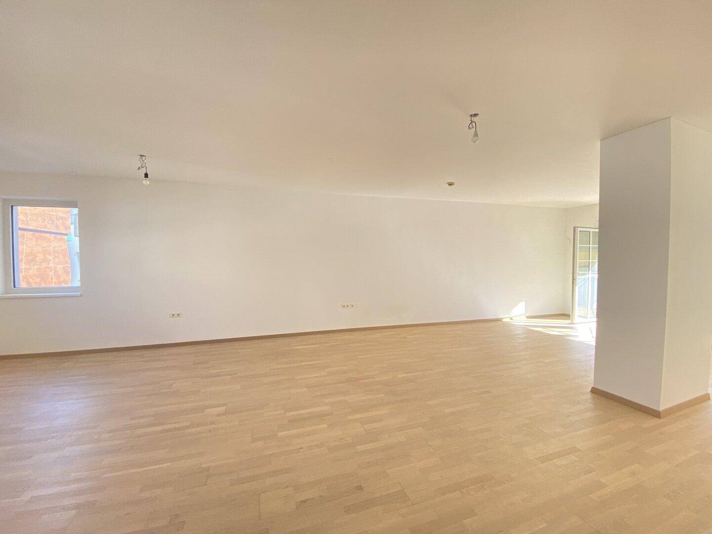 Geräumiger, heller Wohnbereich mit Holzboden und viel Tageslicht durch die Fenster.