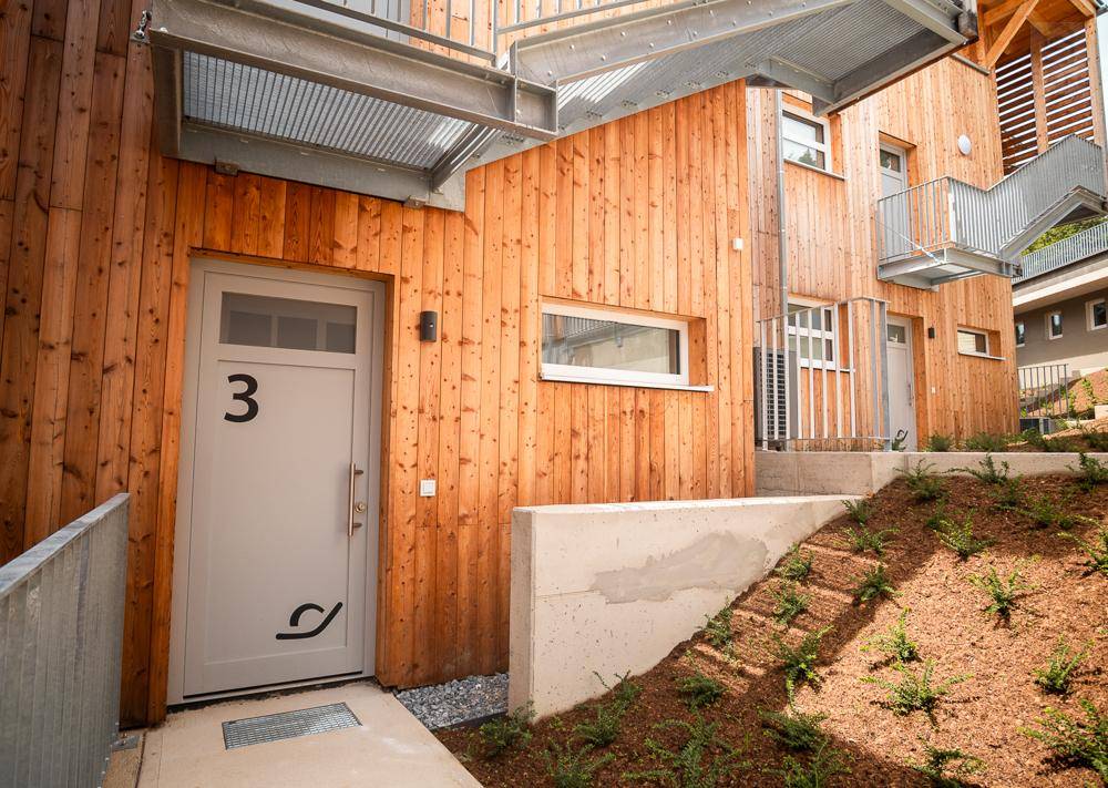 Eingangsbereich einer Wohneinheit mit heller Tür und Holzfassade, umgeben von junger Bepflanzung.