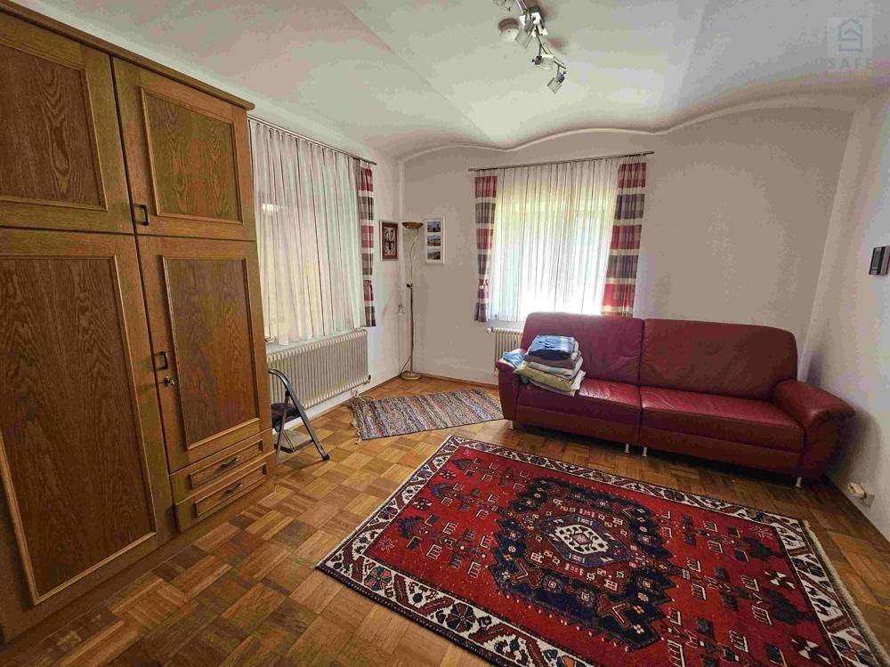 Wohnzimmer mit rotem Sofa, Holzschrank und gemustertem Teppich auf Parkettboden.