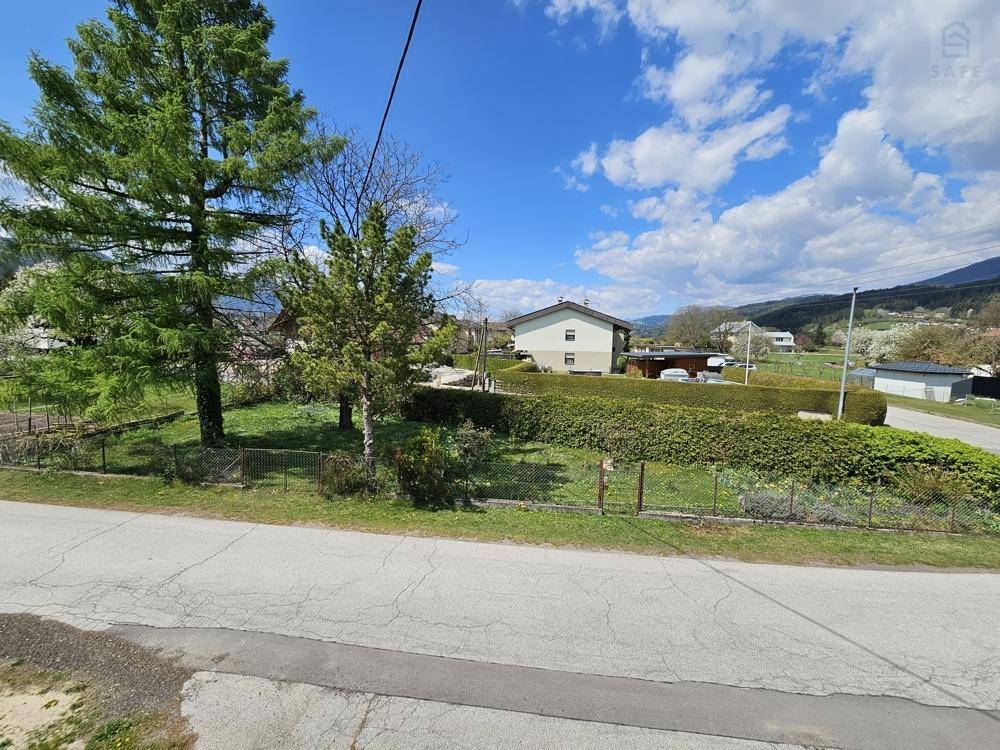 Blick über eine Straße auf eine grüne Hecke und Bäume mit weiteren Häusern im Hintergrund und Bergen.