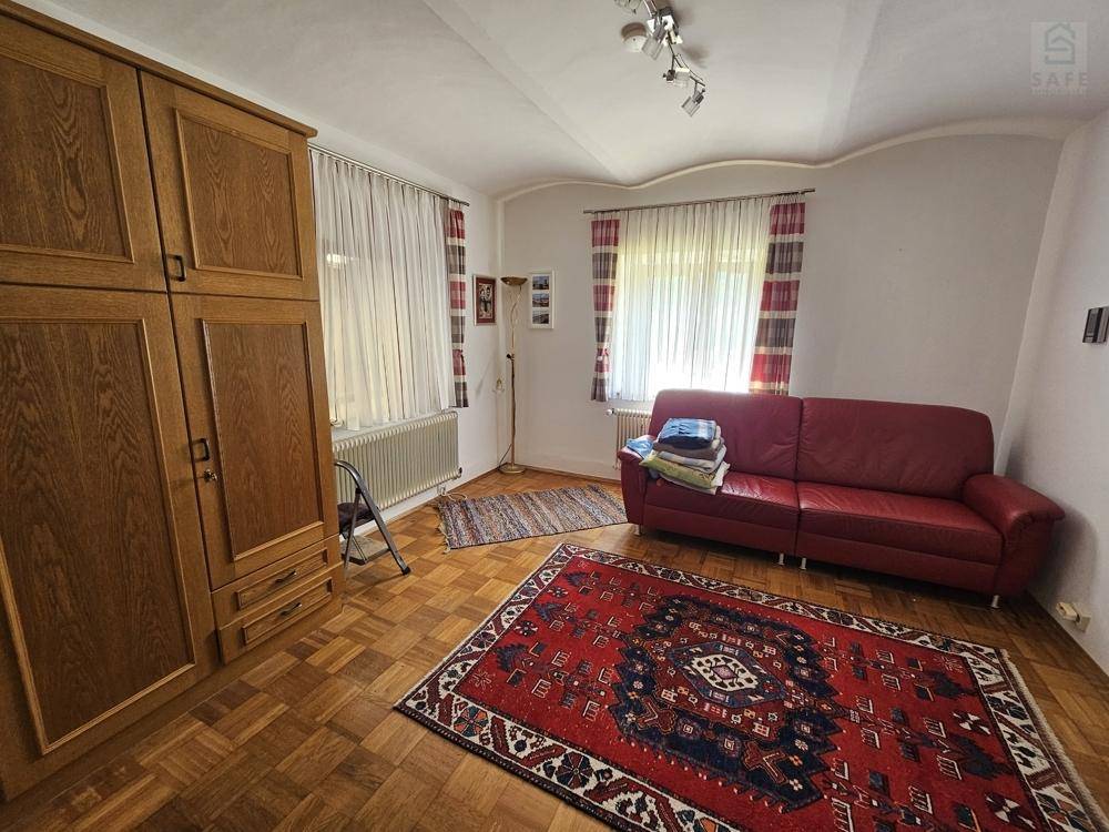 Wohnzimmer mit rotem Sofa, Holzschrank und Parkettboden, beleuchtet durch zwei große Fenster.