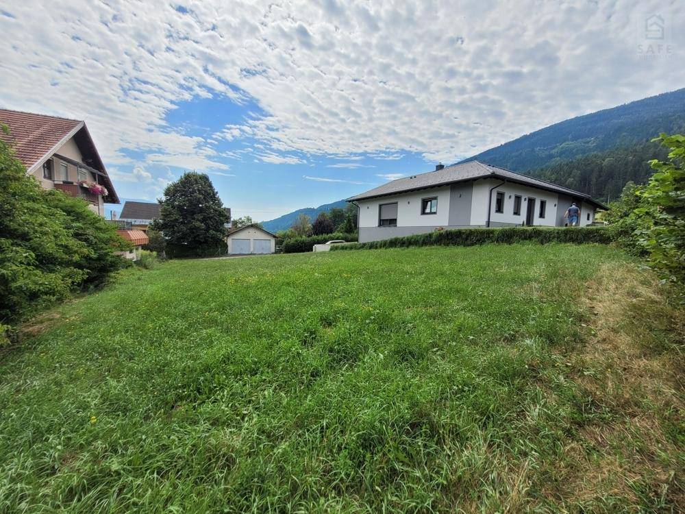 Grünes Grundstück mit einem modernen Einfamilienhaus und Bergblick unter bewölktem Himmel.