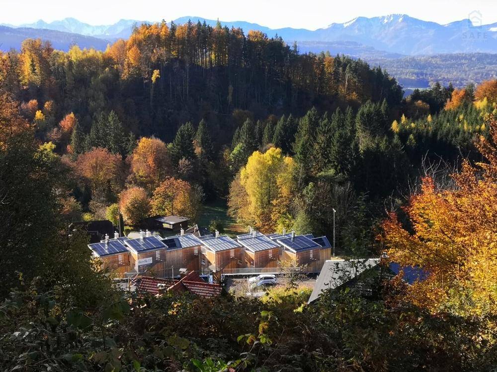 Gruppe moderner Häuser mit Solaranlagen, eingebettet in eine herbstliche Waldlandschaft mit Bergblick.
