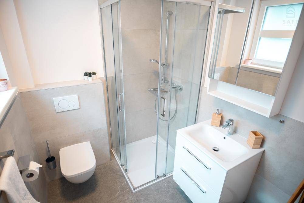 Modernes Badezimmer mit Eckdusche, wandhängendem WC und Fenster, ausgestattet mit grauen Fliesen.