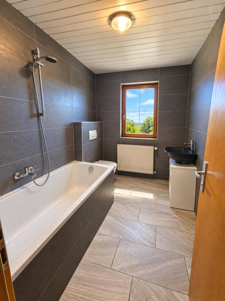 Modernes Badezimmer mit Badewanne, Dusche, Fenster und dunklen Fliesen an Wänden und Boden.