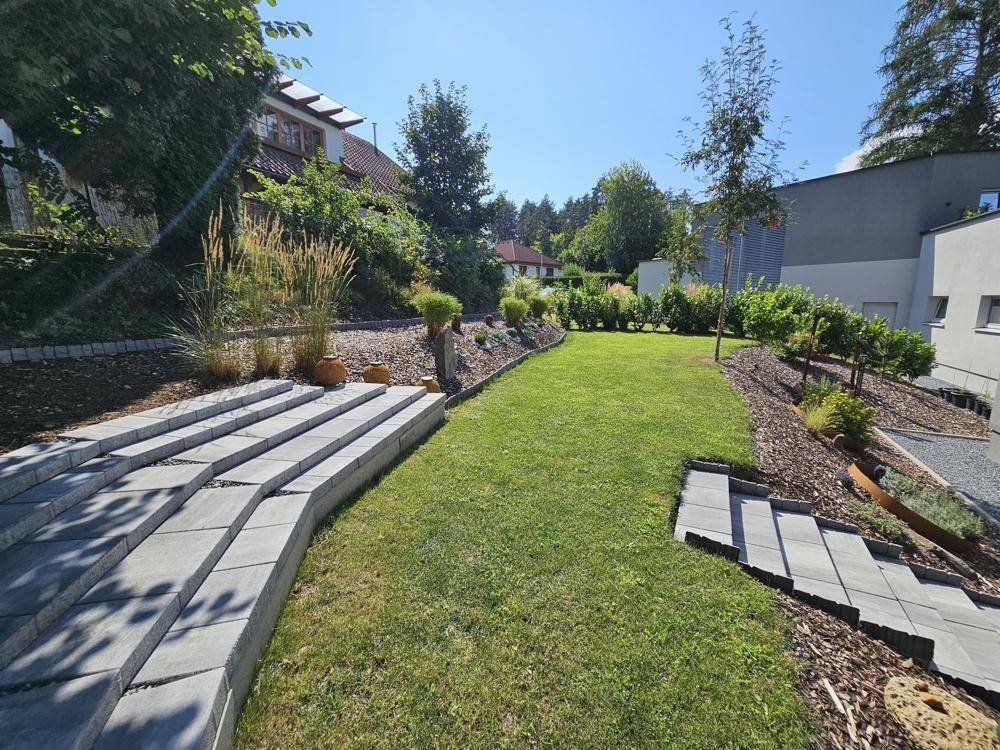 Gestalteter Gartenbereich mit Rasen, Steinstufen und vielfältiger Bepflanzung unter blauem Himmel.