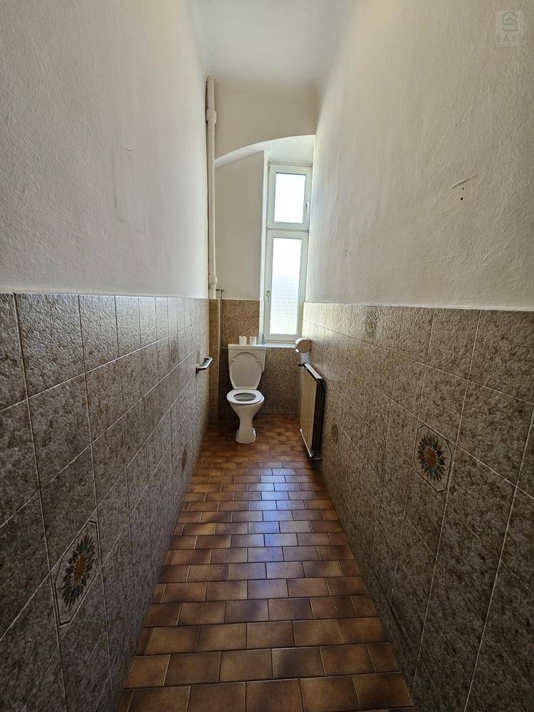 Schmaler Toilettenraum mit braun-beigefarbenen Fliesen an Wänden und Boden sowie einem kleinen Fenster für Tageslicht.