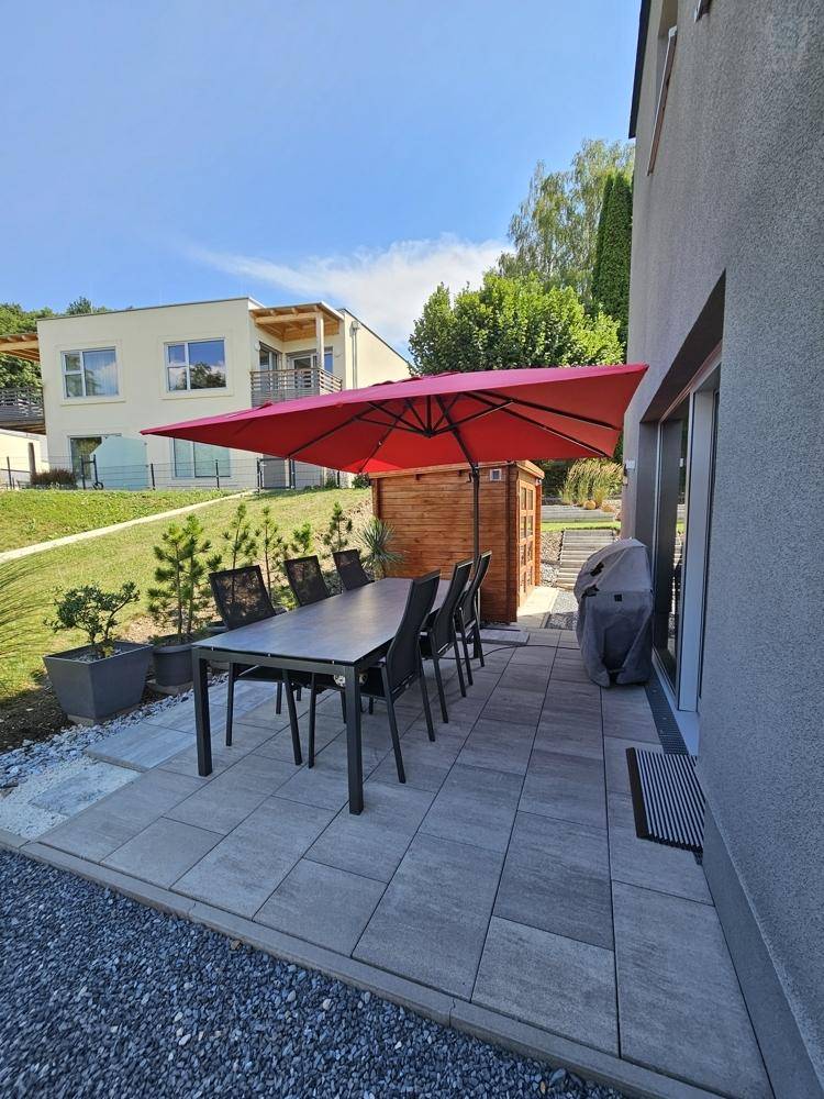 Gepflasterte Terrasse mit großem Esstisch, rotem Sonnenschirm und Grillbereich für Mahlzeiten im Freien.