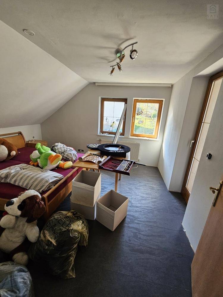 Helles Schlafzimmer im Dachgeschoss mit schrägen Wänden, zwei Fenstern und dunklem Bodenbelag.