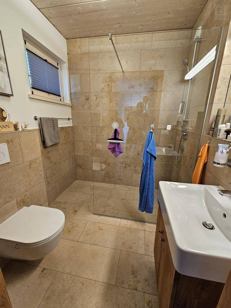 Modernes Badezimmer mit ebenerdiger Dusche, Hänge-WC und hellen Fliesen in Natursteinoptik.