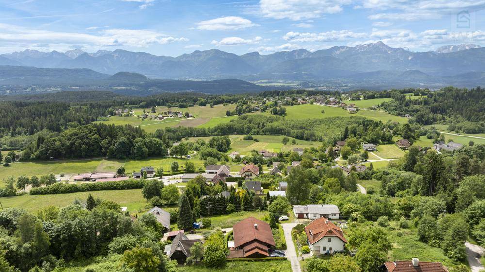 Weitläufige Landschaftsaufnahme mit einem Dorf, Feldern und majestätischen Bergen im Hintergrund unter blauem Himmel.