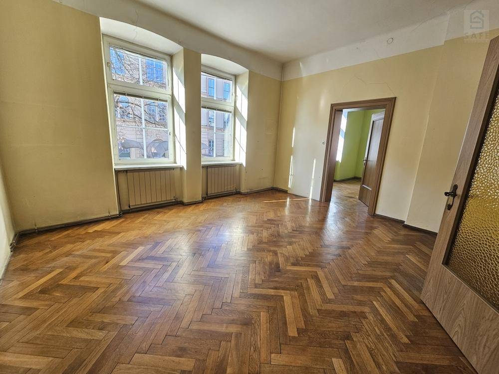 Geräumiges Zimmer mit Fischgrätparkett, hellgelben Wänden und zwei Fenstern, die den Raum mit Tageslicht füllen.
