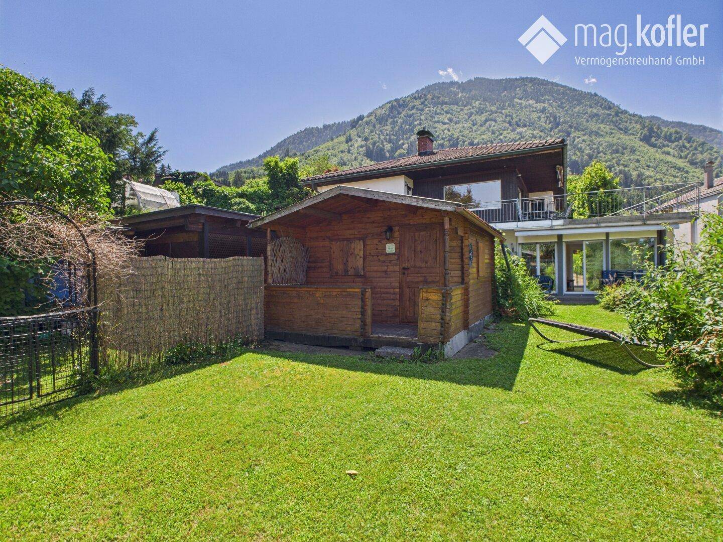 Gemütlicher Holzschuppen im Garten mit dem Haupthaus und Bergen im Hintergrund.