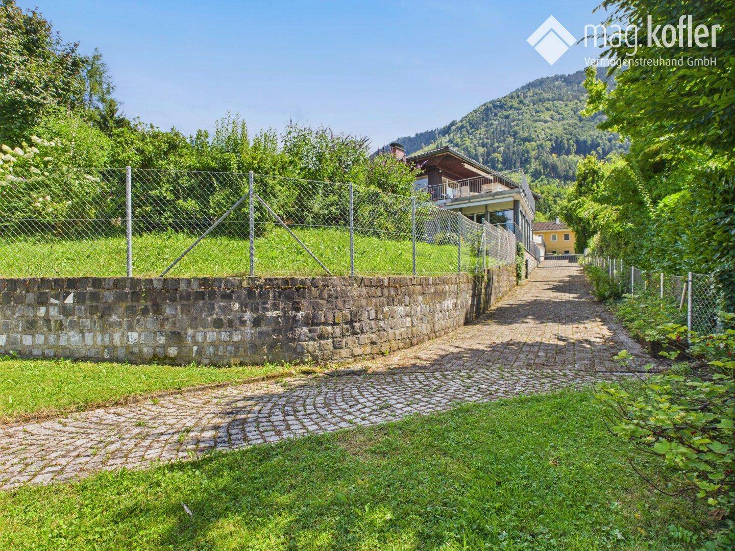 Gepflasterter Zufahrtsweg mit Steinmauer und Blick auf das Haus und die Berge.