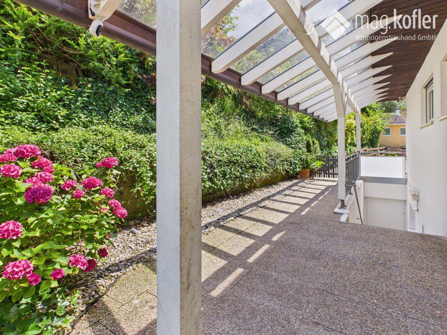 Überdachte Terrasse mit Betonboden und üppiger Bepflanzung entlang einer Mauer.