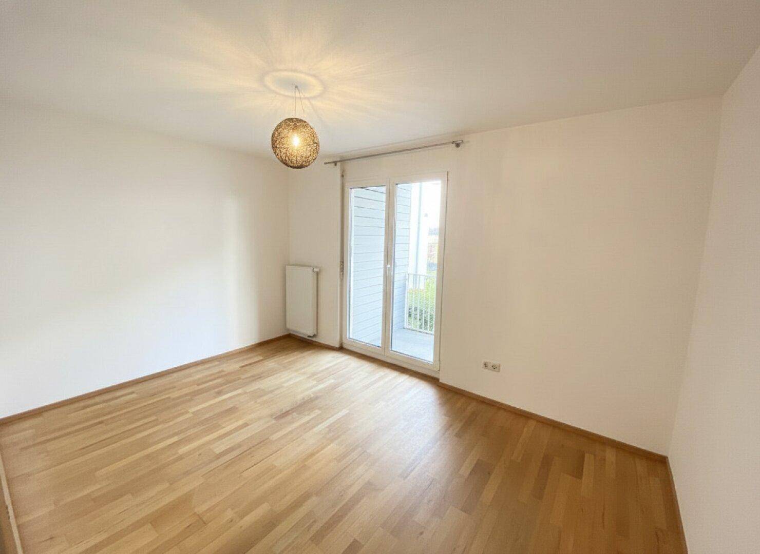 Schlafzimmer mit Holzboden, Heizkörper und Zugang zum Balkon, ausgestattet mit einer modernen Leuchte.