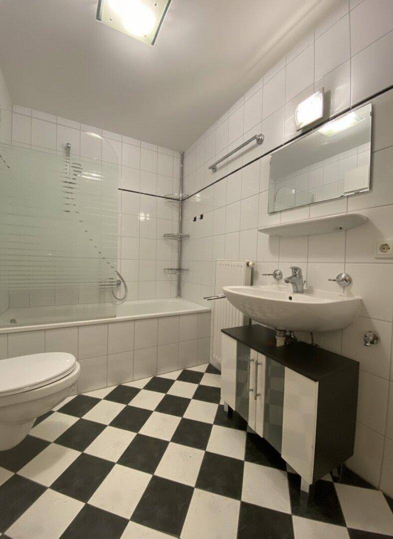 Badezimmer mit Waschbecken, Spiegel und Badewanne mit Duschabtrennung und kariertem Boden.