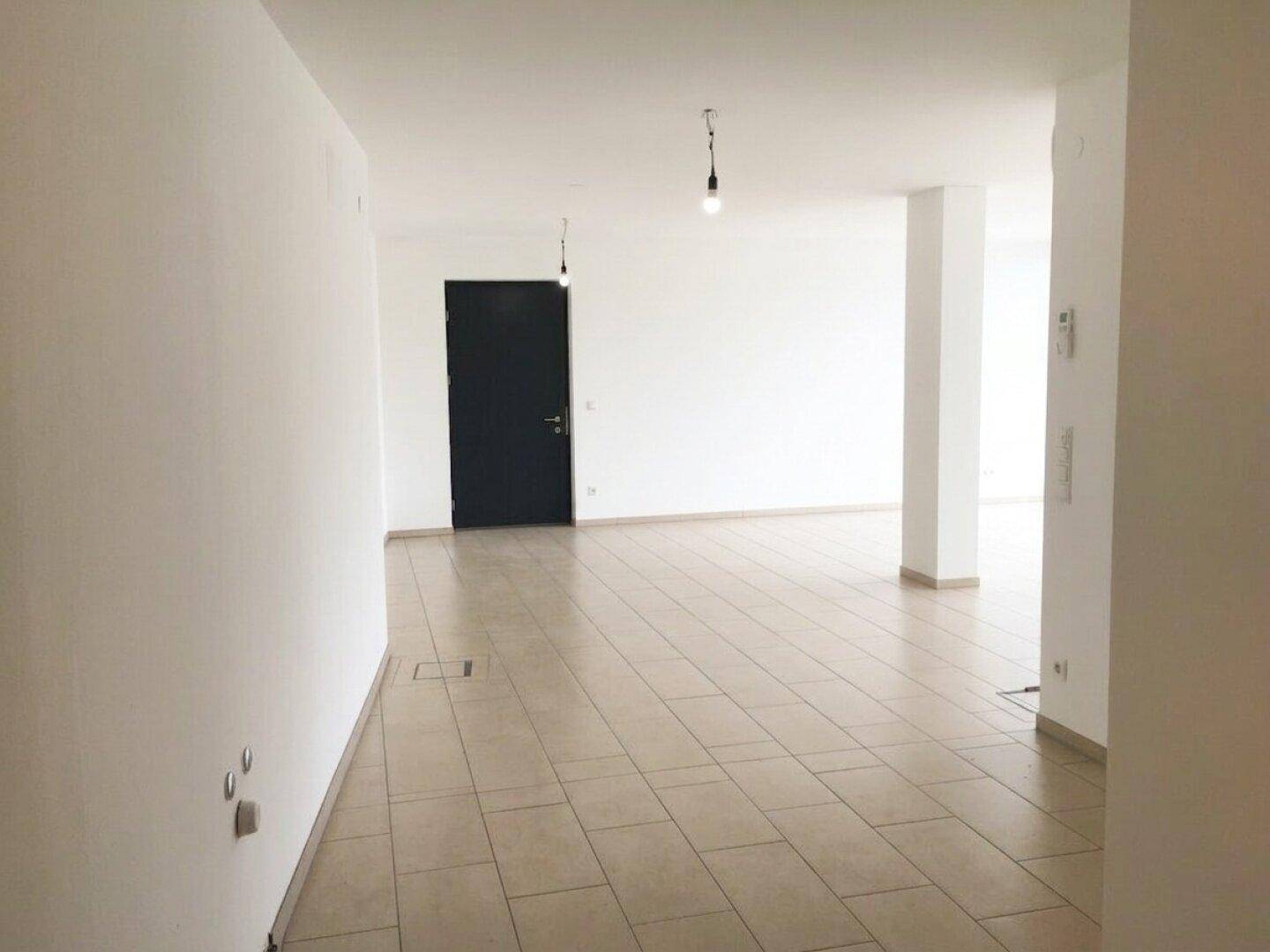 Leerer, heller Raum mit beigefarbenen Fliesen, weißen Wänden und einer dunklen Tür.