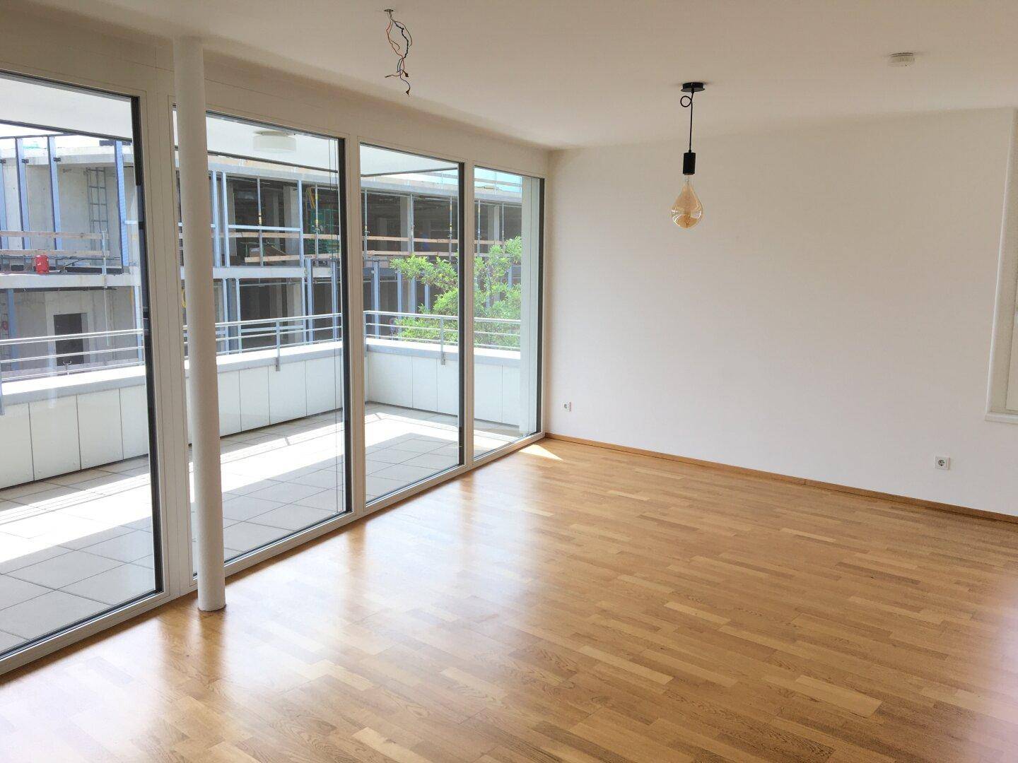 Geräumiges, unmöbliertes Wohnzimmer mit großen Fenstern, Holzboden und Zugang zum Balkon.
