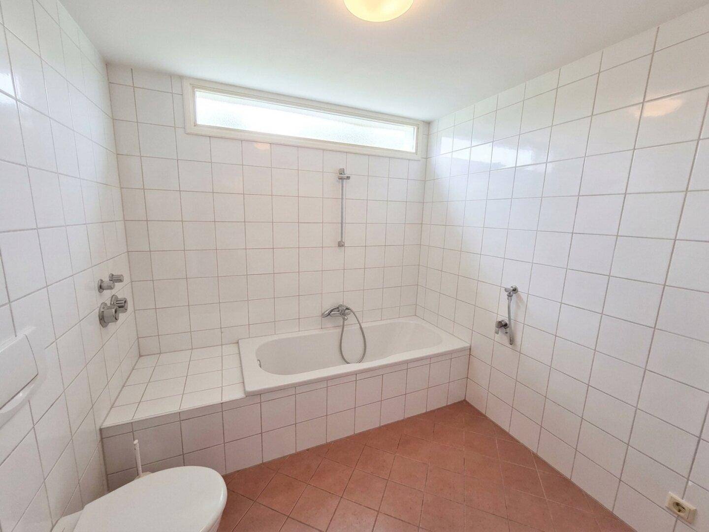 Funktionales Badezimmer mit Badewanne und Toilette, komplett gefliest in Weiß und Rotbraun.