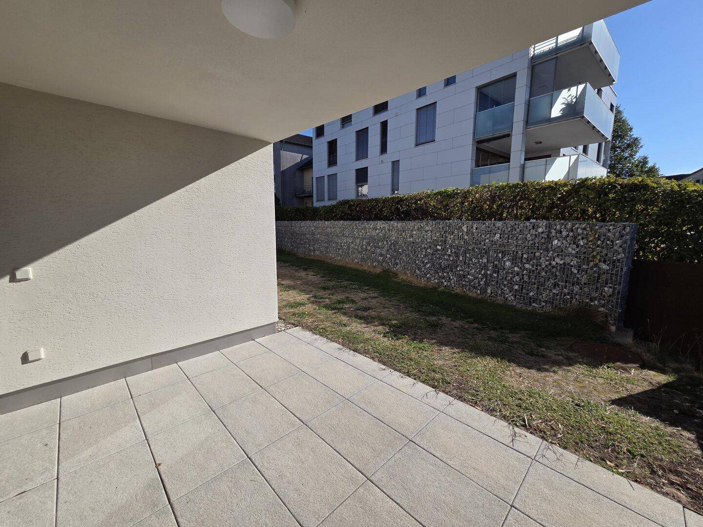 Gepflasterte Terrasse mit kleinem Gartenbereich und Gabionenwand, angrenzend an ein modernes Gebäude.